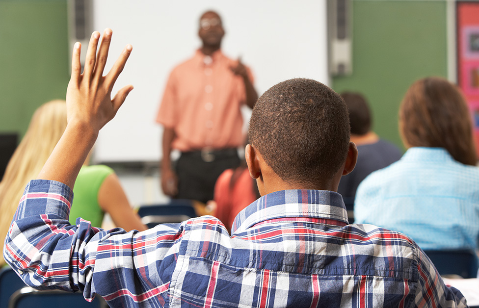 View from behind student in plaid shirt raising hand in front of teacher
